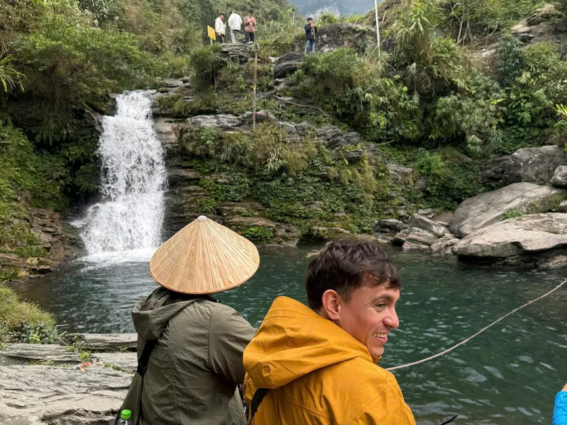 A waterfall dropping into a green pool in Du Gia, travellers sitting on the rocks beside it