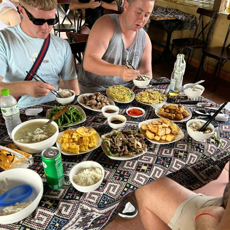 A table of shared dishes at a homestay, travellers eating with chopsticks on a woven cloth