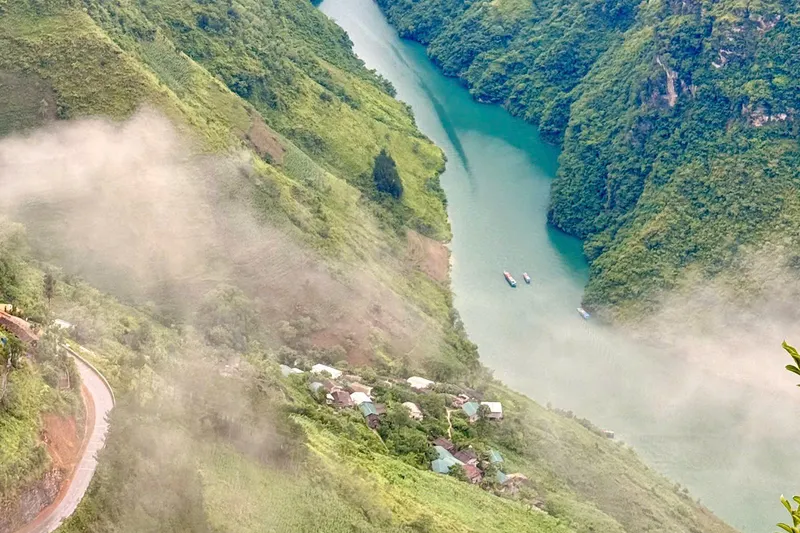 An aerial view of the Nho Que river cutting through the limestone gorge beneath Ma Pi Leng, boats on the water and a village at the foot of the cliff
