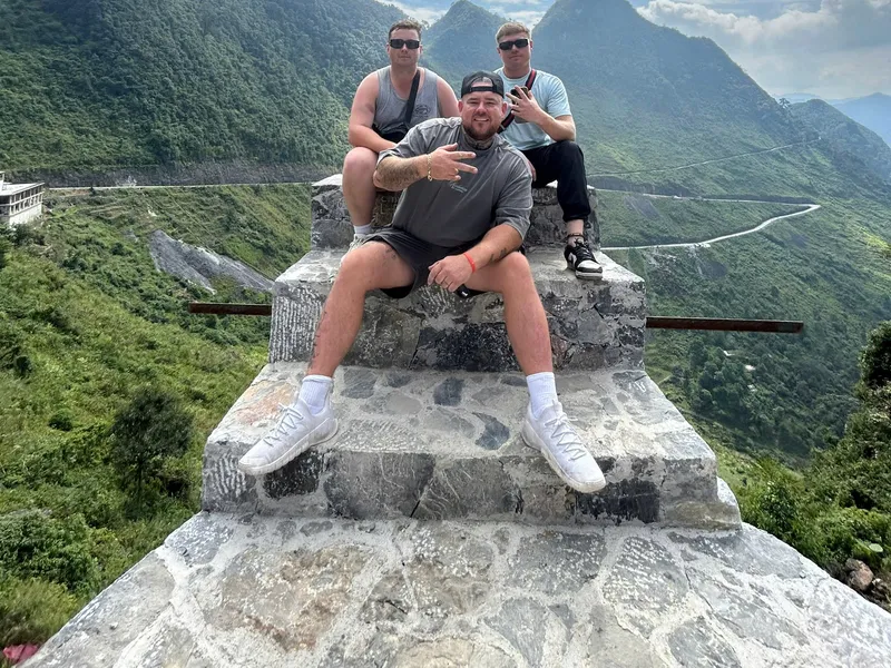 Three travellers sitting on the stone viewpoint platform at Ma Pi Leng, the winding pass road visible in the valley below