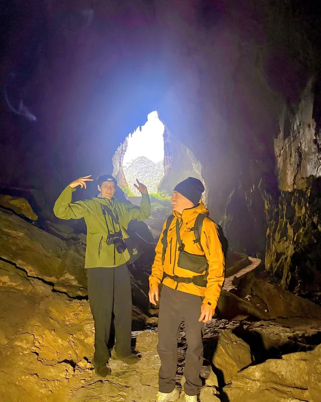 Two travellers with headlamps inside Nguom Ngao Cave, daylight breaking through a narrow opening in the rock