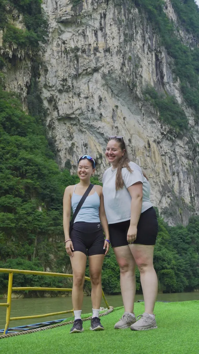 Two travellers celebrating on a boat raft beneath a towering limestone cliff on the Nho Que river