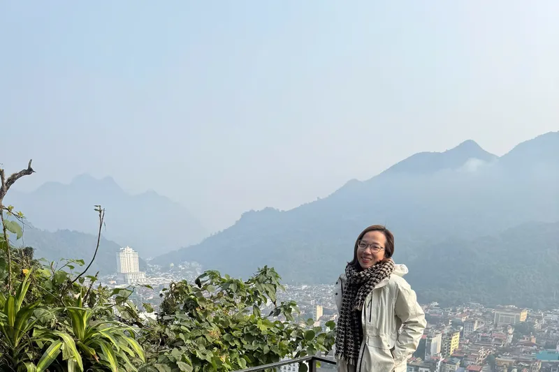 A traveller smiling on a rooftop deck, a Ha Giang town spread out in the valley behind her