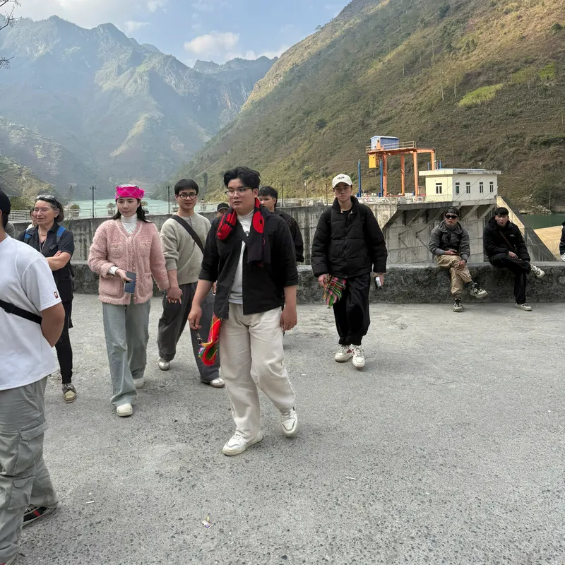 A small group of travellers walking across a paved viewing platform above a valley, railings and a dam visible behind them