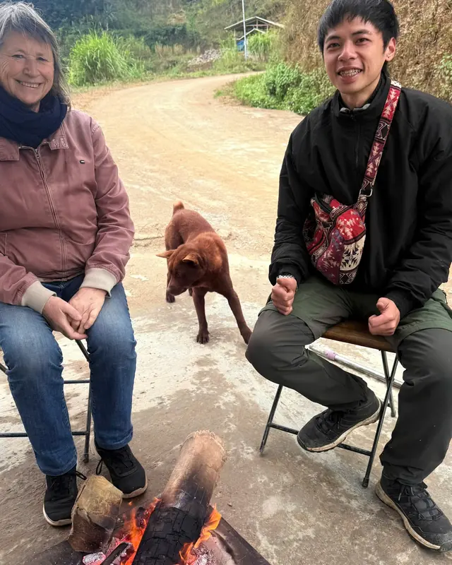 Matt sitting by a roadside fire with a traveller, a dog crossing the dirt track behind them